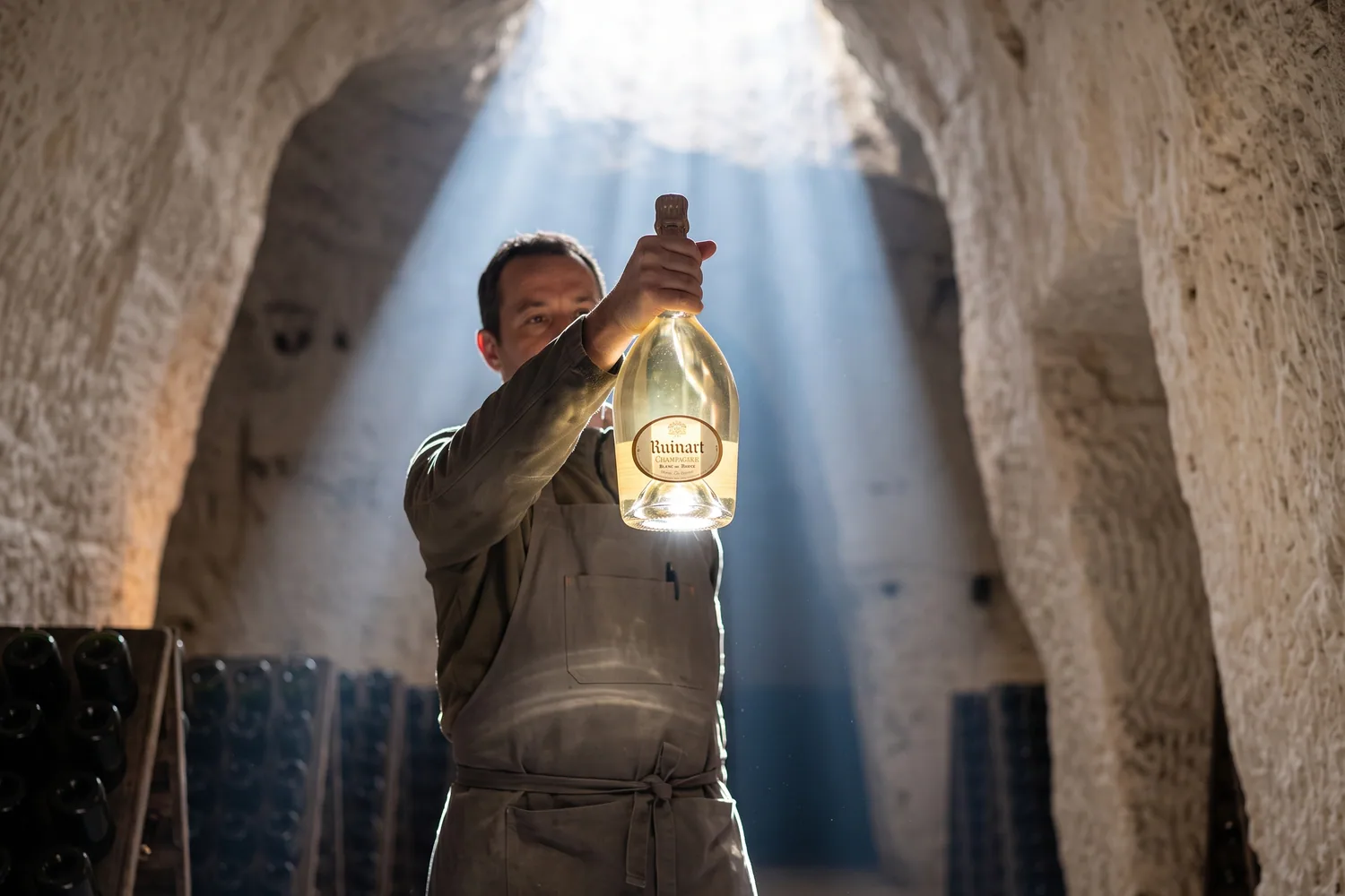 Caviste levant une bouteille de Ruinart Blanc de Blancs dans un rai de lumière, au cœur des crayères de Reims